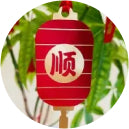 Red paper lantern with white stripes and Chinese character, hanging among green leaves. Conveys a festive, traditional atmosphere.