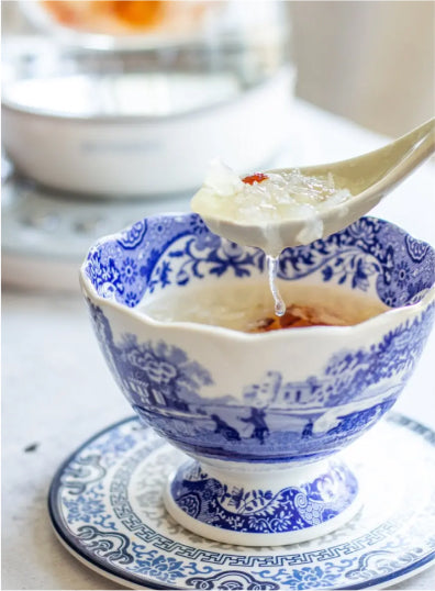 Traditional Chinese blue and white porcelain bowl filled with bird's nest soup, a spoon lifting up a portion highlighting the delicate texture and a goji berry, presented on a matching saucer with soft natural lighting.