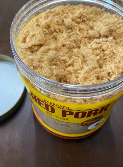 Open jar of seasoned dried pork floss with fluffy, golden-brown texture. The label is partially visible. Lid rests on a wooden surface nearby.
