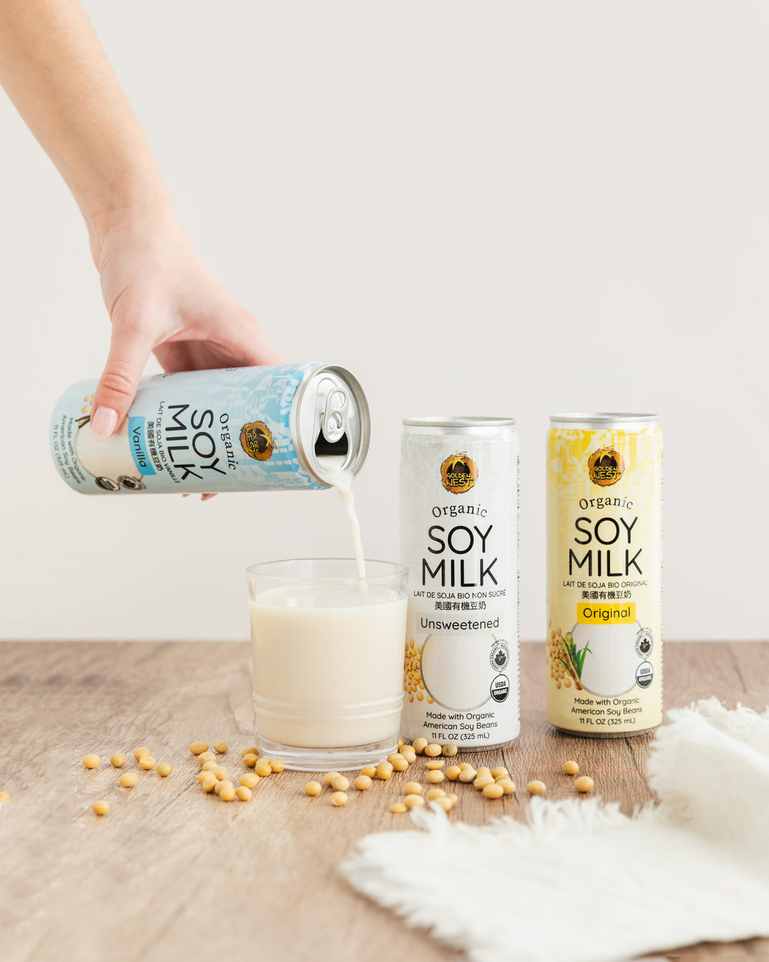 A hand pours vanilla organic soy milk from a blue can into a glass. Two other cans, labeled original and unsweetened, stand nearby on a wooden table.
