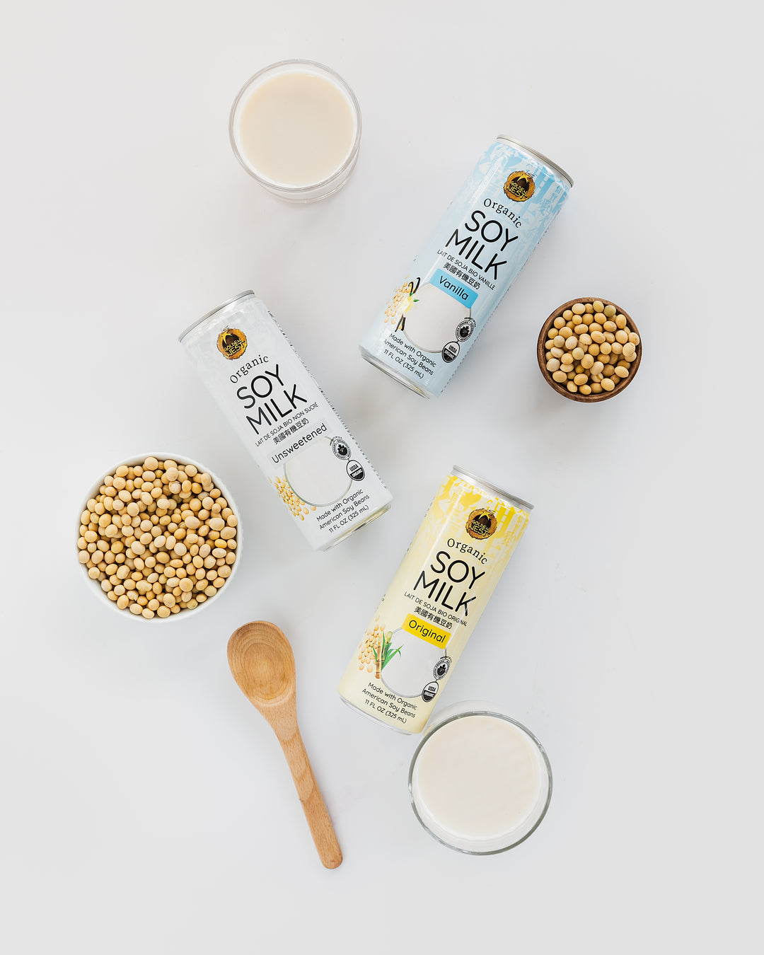 Three cans of organic soy milk labeled Unsweetened, Vanilla, and Original are arranged with glasses of milk, bowls of soybeans, and a wooden spoon on a white background.