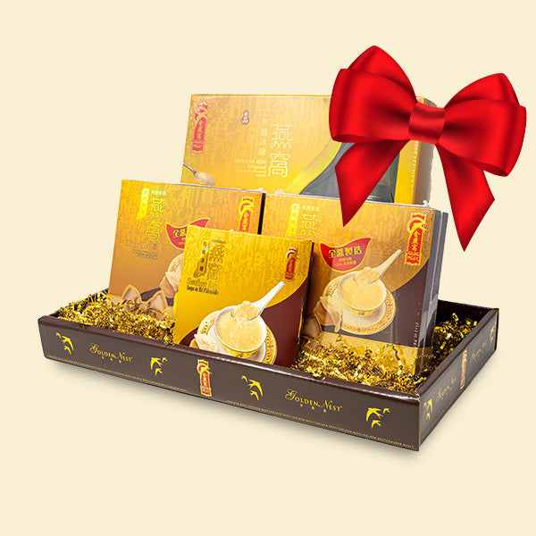 Gift box with various yellow and brown packaged items, accented with shiny gold filling. A red bow sits on top. Elegant and festive presentation.