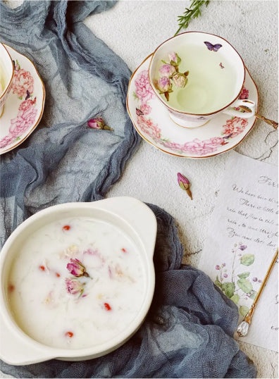 A serene tea setting with floral teacups, green tea, pink accents, blue cloth, a bowl of rose petal oatmeal, and a handwritten note creating an elegant, peaceful mood.