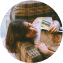A person with long hair, resting on a plaid couch with eyes closed, holds an open magazine. The scene feels peaceful and relaxed.