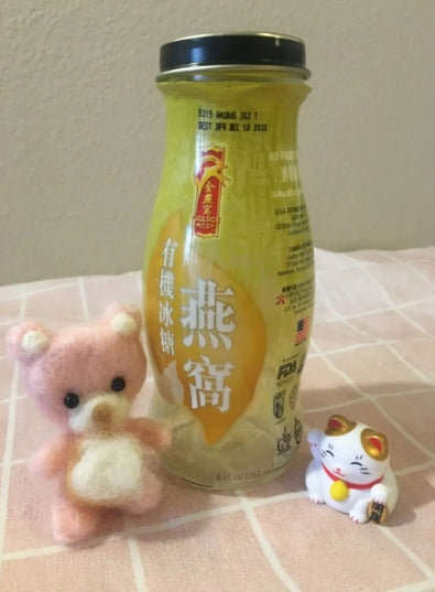 在粉紅色格子的表面上,一瓶帶有黑色蓋子的中國燕窩飲料坐落在一個粉紅色的小泰迪熊人像和一隻陶瓷 Maneki-neko 貓之間。
